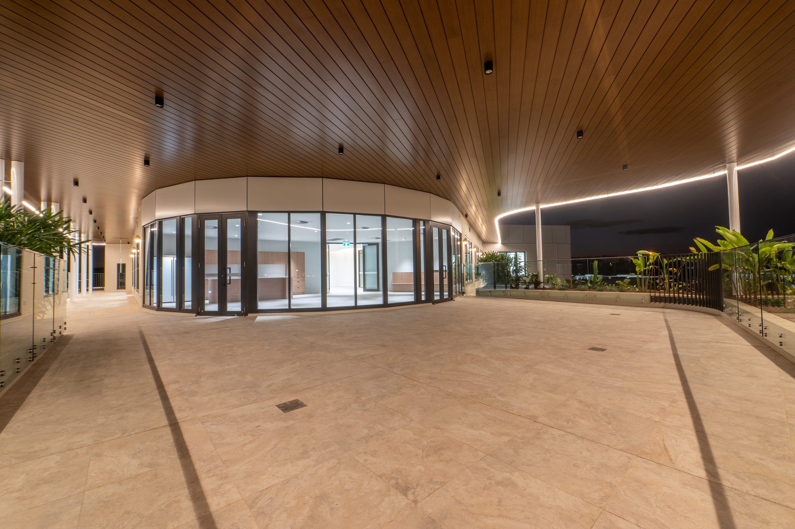 Covered walkway with a timber-lined ceiling of downlights and continuous linear LED lighting at dusk
