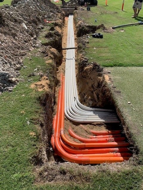 Open trench with bundled orange and grey electrical conduits laid through a sports field