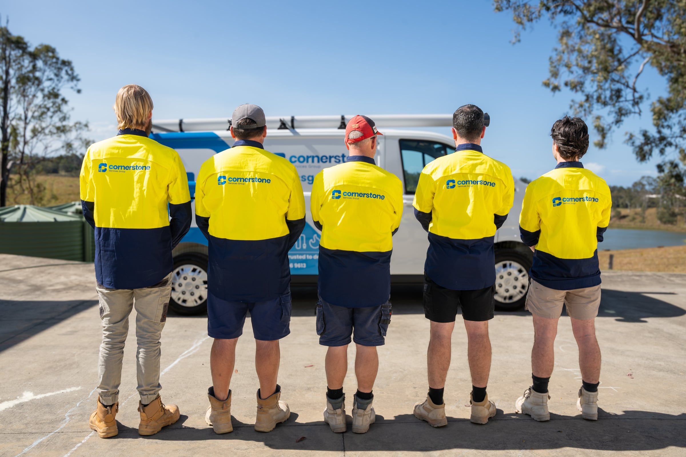 Five Cornerstone Services Group team members seen from behind, branded work shirts facing a company van